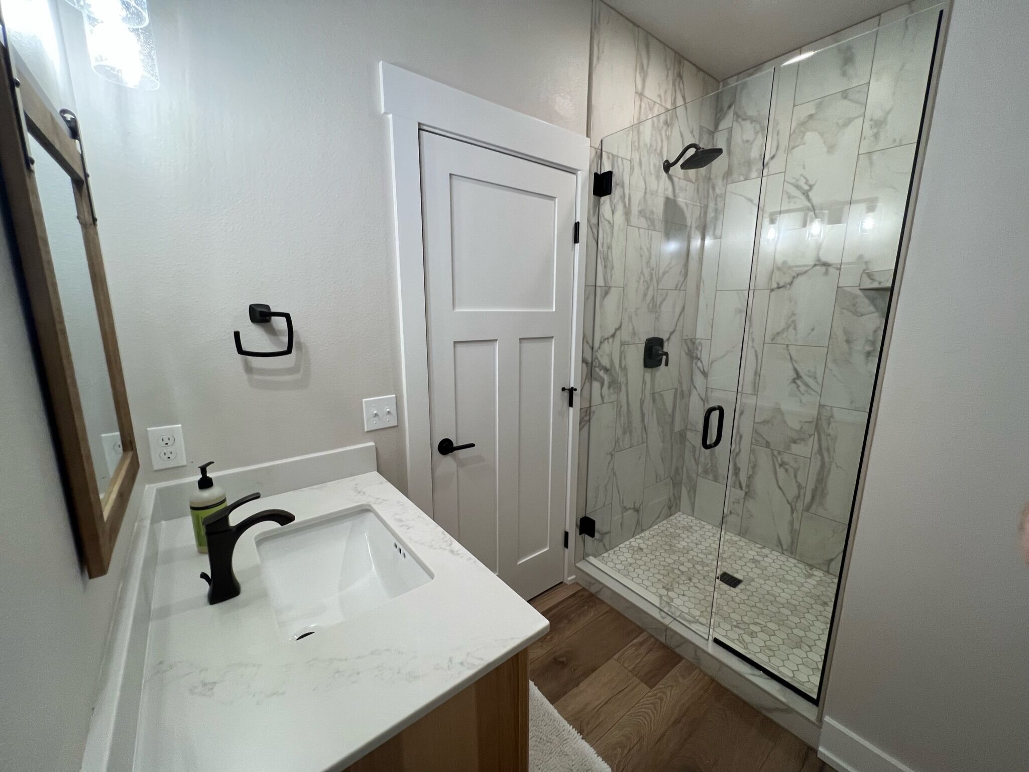 Un baño con un lavamanos blanco, un espejo, una ducha con puertas de vidrio y una puerta blanca.