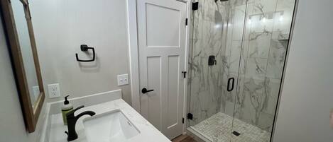 Un baño con un lavamanos blanco, un espejo, una ducha con puertas de vidrio y una puerta blanca.