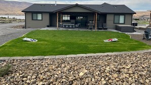 A modern house with a covered patio, a lawn, and a rocky area in the foreground. - New Custom Multi Master + Sleeping Room Golf Course Home w/ Outdoor Living Area  (Mattawa)
