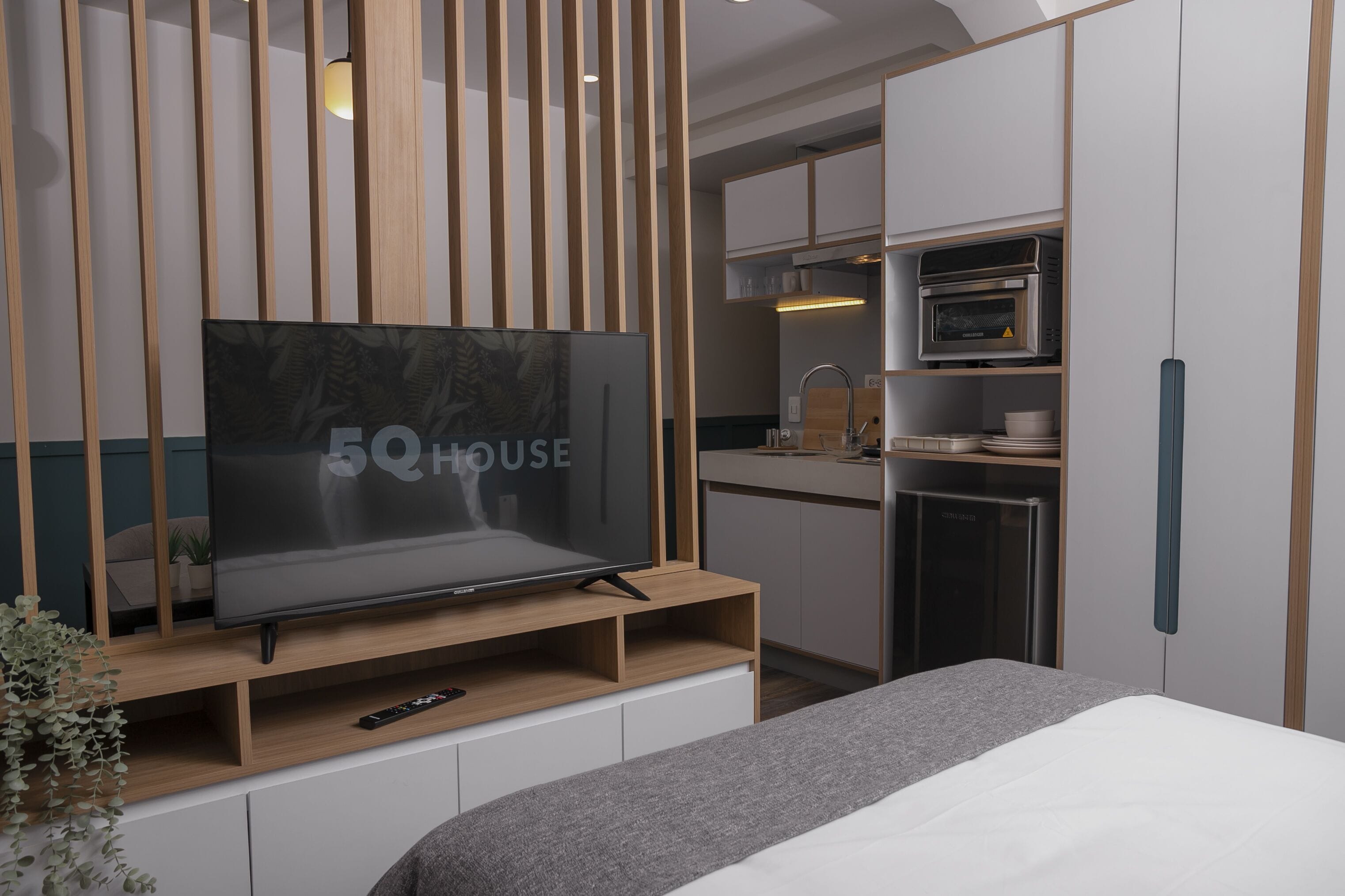 A modern hotel room with a TV on a wooden stand, a kitchenette with a microwave, and a bed with a grey blanket.