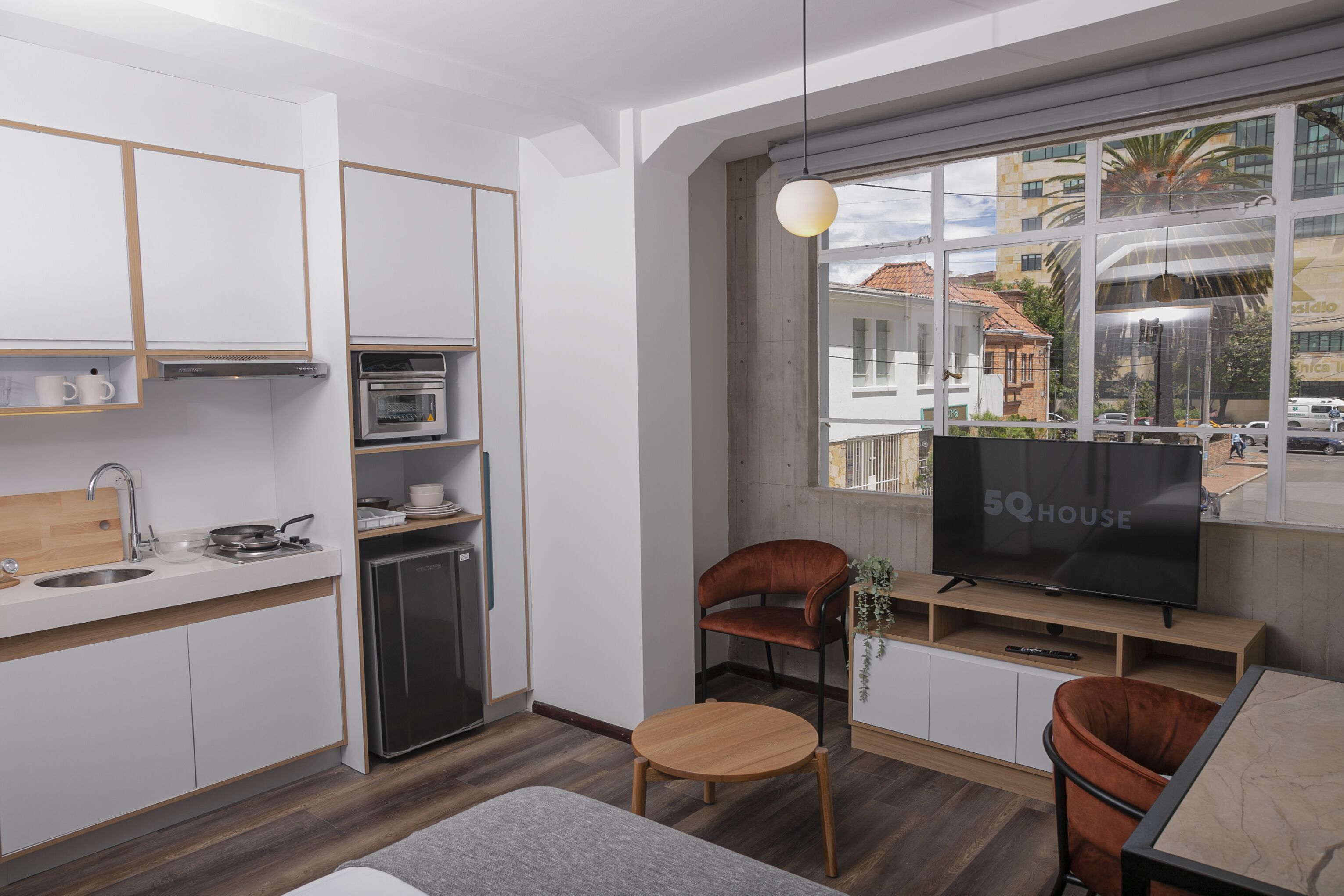 A modern kitchen with white cabinets, a built-in oven, and a small refrigerator. There is a wooden dining table with two chairs, a TV on a wooden stand, and a window with a view of buildings and cars outside.