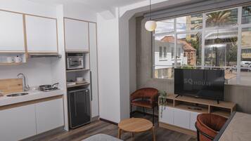 A modern kitchen with white cabinets, a built-in oven, and a small refrigerator. There is a wooden dining table with two chairs, a TV on a wooden stand, and a window with a view of buildings and cars outside.