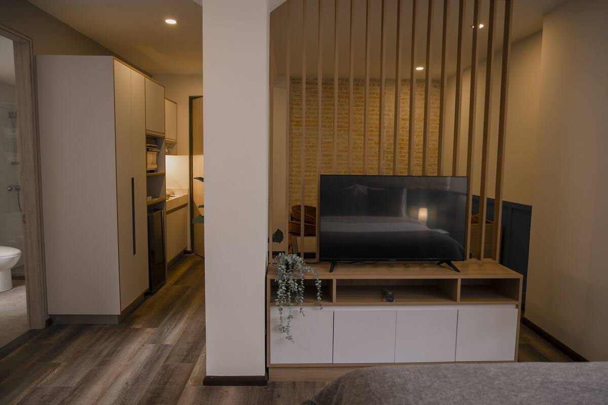 A modern bedroom with a TV on a wooden stand, a bed, and a small kitchenette in the background.