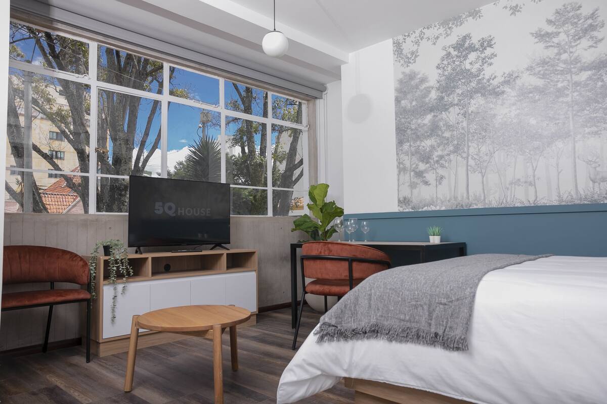 A modern hotel room with a large window, a flat-screen TV, a sofa, a bed, a small table, and a desk.
