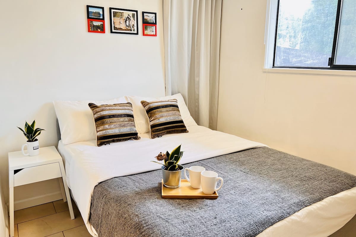 Ein ordentlich bezogenes Bett mit gestreiften Kissen, ein Nachttisch mit Pflanze und ein Fenster mit Vorhängen.