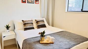 Una cama bien hecha con almohadillas a rayas, una mesita de noche con una planta y una ventana con cortinas.