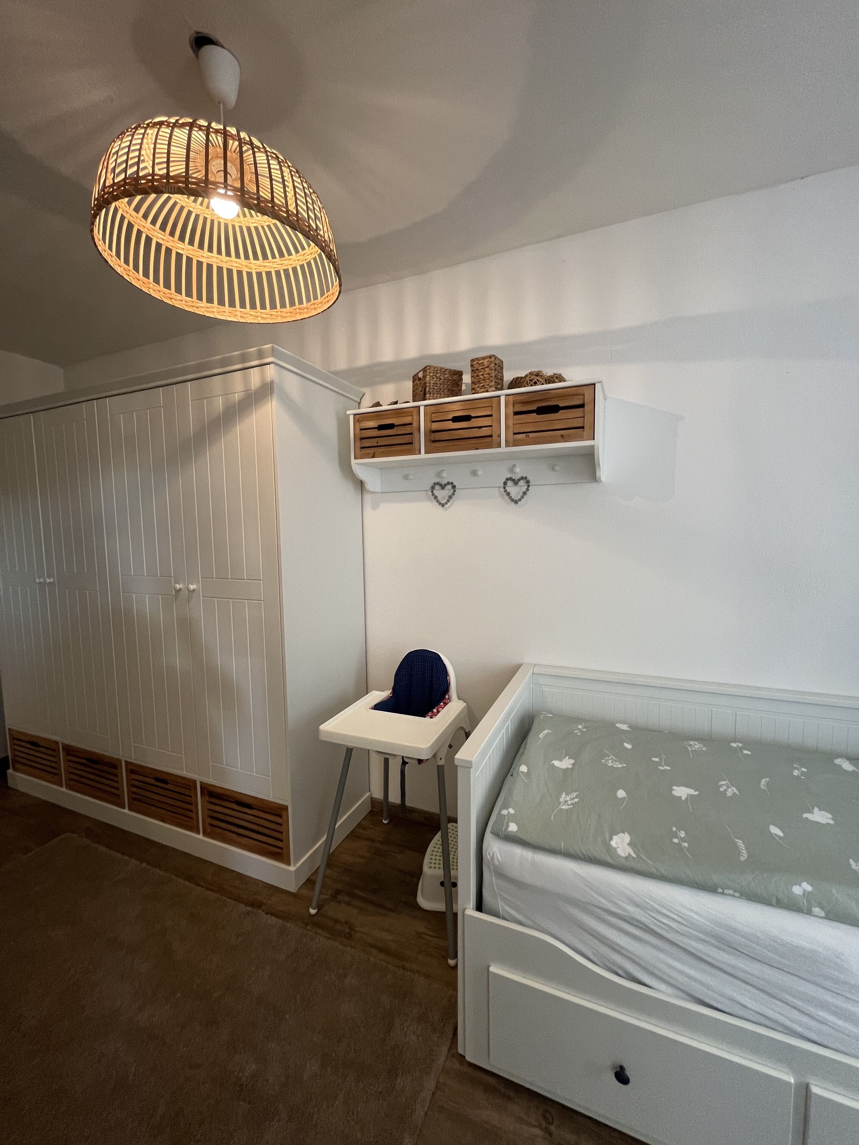 A child's room with a bed, a high chair, a wardrobe, and a shelf with storage boxes and heart-shaped decorations.