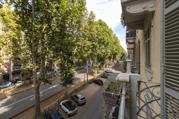 A street view with parked cars, trees lining the road, and buildings with balconies.
