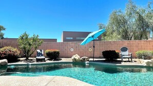 Una piscina azul cristalina con una sombrilla azul, rodeada por un muro de ladrillo, y una silla.