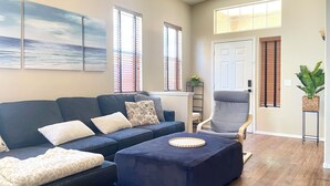 Una sala de estar con un sofá azul oscuro, un reposapiés a juego y un sillón gris. Hay tres ventanas con persianas, una puerta blanca y una planta sobre una mesita auxiliar.