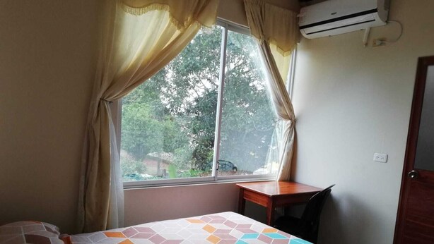 A bedroom with a bed, a desk, and a window with curtains. - Hostal Amazónico Bello Amanecer (Nueva Loja)