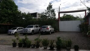 A parking area with three cars parked in front of a building with a red roof and white walls. There are several potted plants in the foreground. - Hostal Amazónico Bello Amanecer (Nueva Loja)