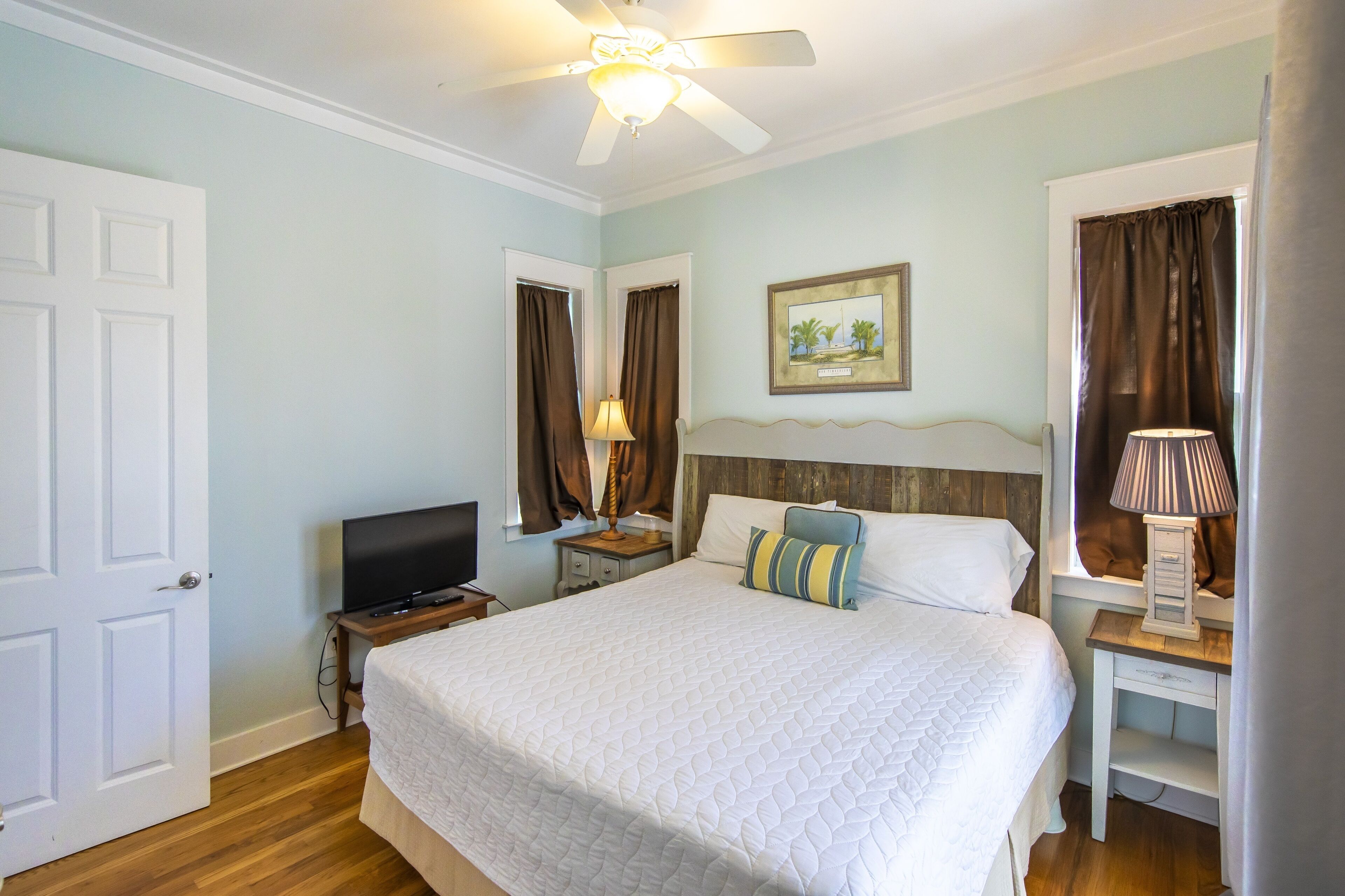 Quarto bem arrumado com cama, televisão, ventoinha no teto e mesa de cabeceira com abajur.