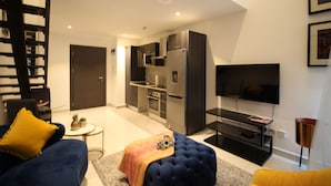 A modern living room with a sofa, ottoman, and a flat-screen TV. - Embassy Gardens - One Bed (Accra)
