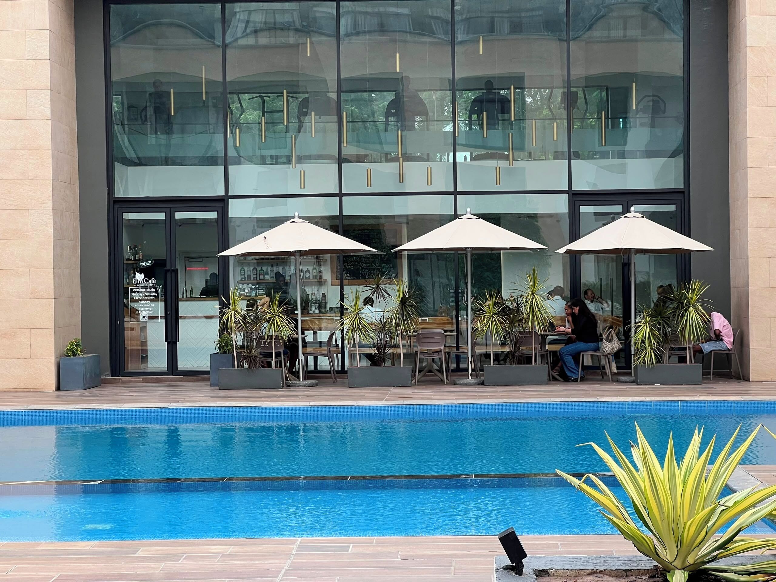 A modern building with a glass facade, outdoor seating with umbrellas, and a pool area.