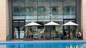 A modern building with a glass facade, outdoor seating with umbrellas, and a pool area. - Embassy Gardens - One Bed (Accra)