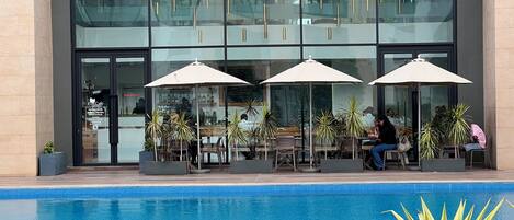 A modern building with a glass facade, outdoor seating with umbrellas, and a pool area.