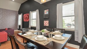 A dining area with a dark wooden table set for four, surrounded by chairs. There is a sofa with a red cushion in the background, and a window with white curtains. - Love&Mystique Getaway in Galleria w/ Pool //Sleeps12! (Houston)