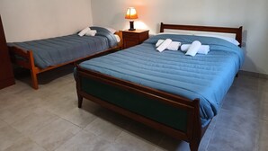 Deux lits simples avec des cadres en bois et une literie bleue, une table de chevet avec une lampe et un sol carrelé.