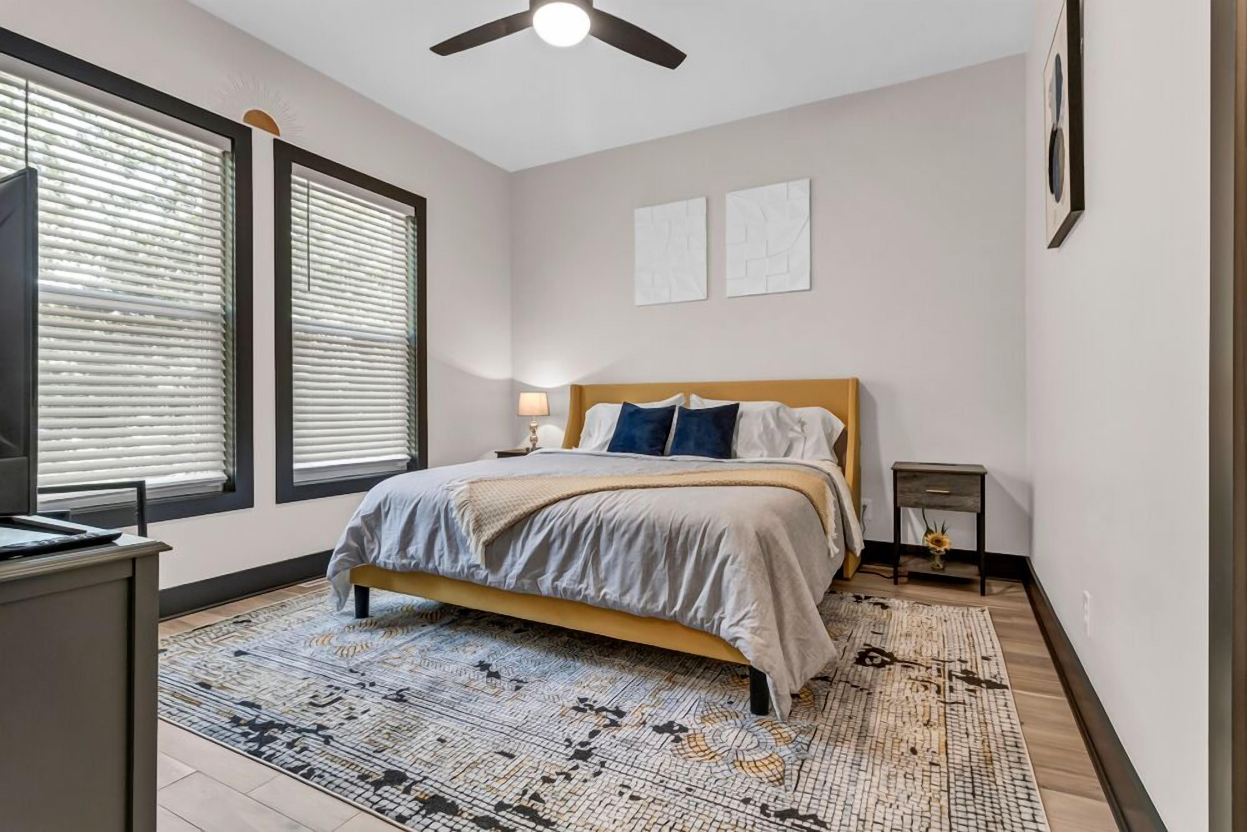 A bedroom with a bed, a desk, a chair, a standing lamp, and a window with blinds.