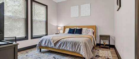 A bedroom with a bed, a desk, a chair, a standing lamp, and a window with blinds.