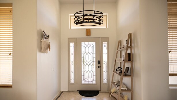 A hallway with a modern chandelier, a decorative door with stained glass, and a wooden ladder shelf with various items. - Desert Ranch House (Page)