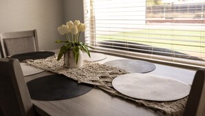 A dining area with a table set for two, a vase with white flowers, and a view through a window with blinds. - Desert Ranch House (Page)