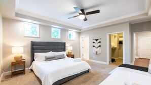 Un dormitorio con una cama grande, dos mesitas de noche con lámparas, un ventilador de techo y vistas a un baño a través de una puerta abierta.