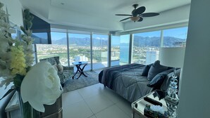 A modern bedroom with a large bed, a ceiling fan, a small table, and a view of a cityscape through large windows.