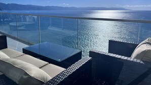A balcony with a glass railing, outdoor furniture, and a view of the sea and distant mountains.