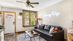 A living room with a black leather sofa, a wooden coffee table, a white door, a window with curtains, and a neon sign on the wall. - Stylish Uptown Butte Condo - 632 (Butte)
