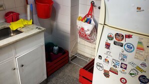 A compact kitchen with white cabinets, a red bucket, a white refrigerator with various magnets, and a red trash bin. - 2 ambientes zona facultad de medicina (Buenos Aires)