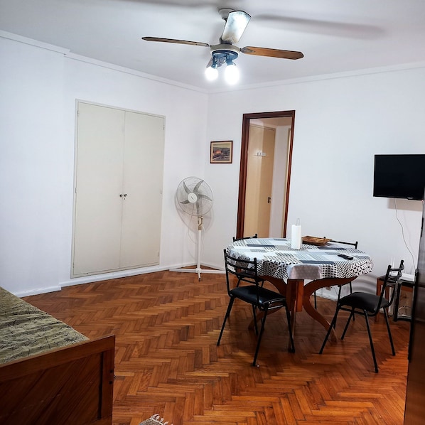 A room with a wooden herringbone floor, a ceiling fan, a dining table with chairs, a TV, and a wardrobe.