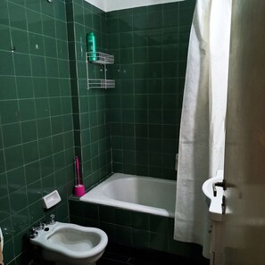 A small bathroom with green tiles, a white bathtub, a white sink, and a pink cleaning brush.