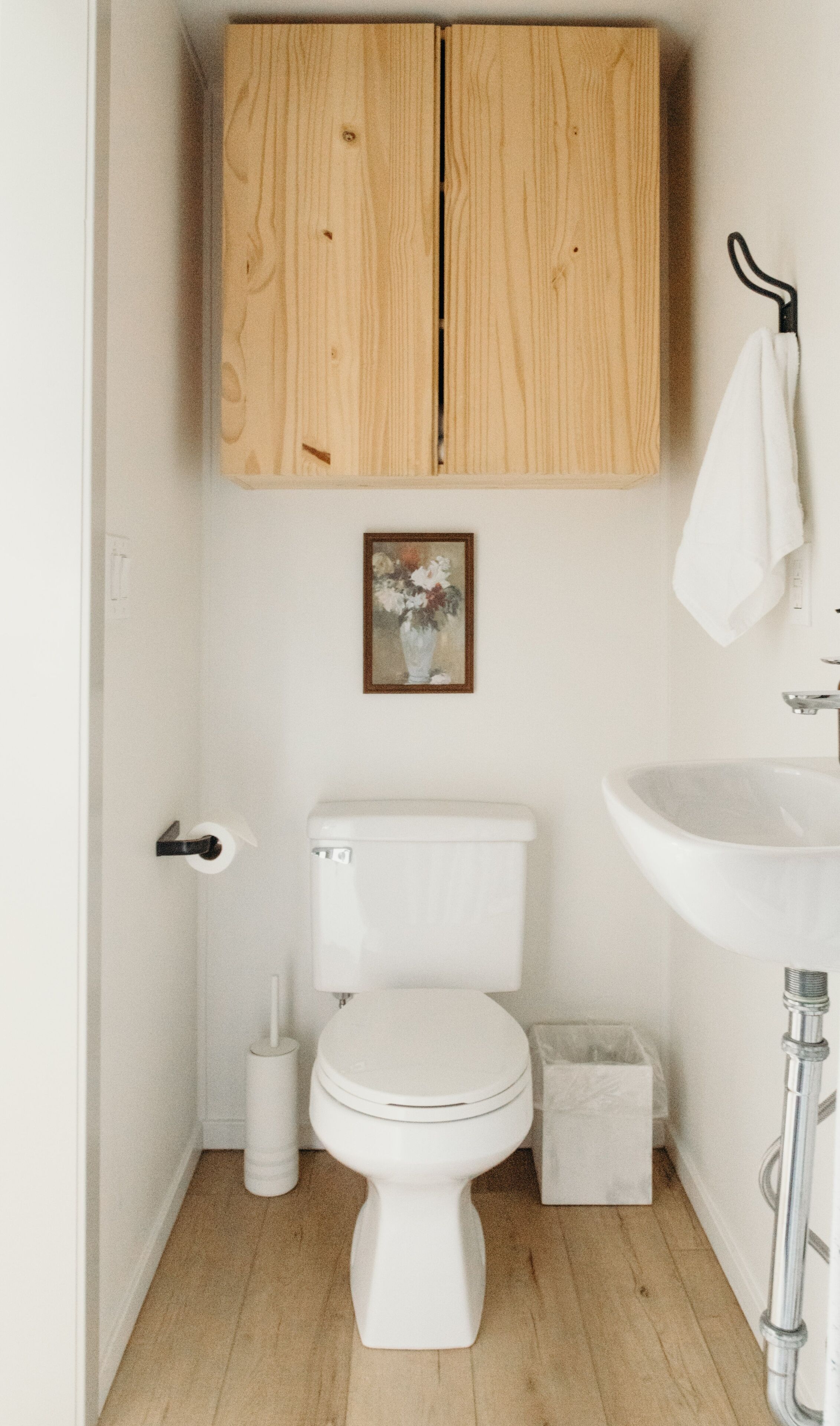 Un baño pequeño con un inodoro blanco, armarios de madera, un lavamanos y un cuadro en la pared.