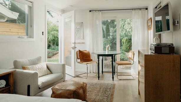 Ein moderner, gut beleuchteter Raum mit einer Couch, einem kleinen Tisch und einem Sessel. Ein Fenster bietet Blick auf Grünflächen.