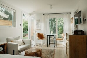 Une pièce moderne et bien éclairée, avec un canapé, une petite table et une chaise. Une fenêtre offre une vue sur la verdure à l’extérieur.