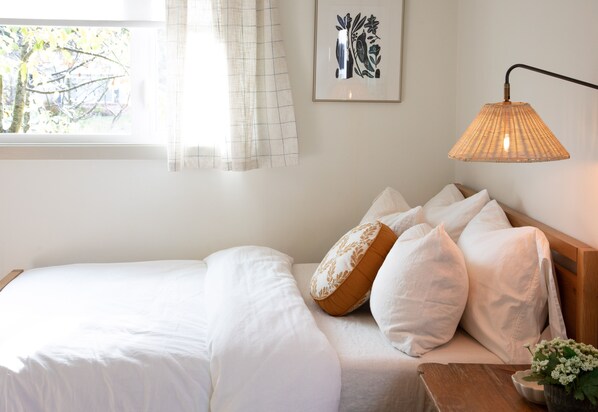Un lit bien fait, avec du linge de lit blanc et plusieurs oreillers, une table de chevet avec une lampe, et une œuvre d’art encadrée au mur.