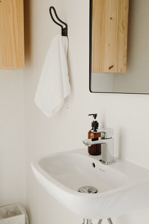 Un lavabo blanc dans une salle de bain, avec un distributeur de savon, une serviette suspendue à un crochet noir et un meuble en bois.