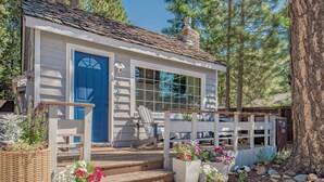 A small, single-story house with a blue door, white siding, and a stone chimney. There are steps leading to a porch with a bench and potted plants. - Steps From the Beach. Tahoe Tipsy Trout. It's a VIBE (Kings Beach)