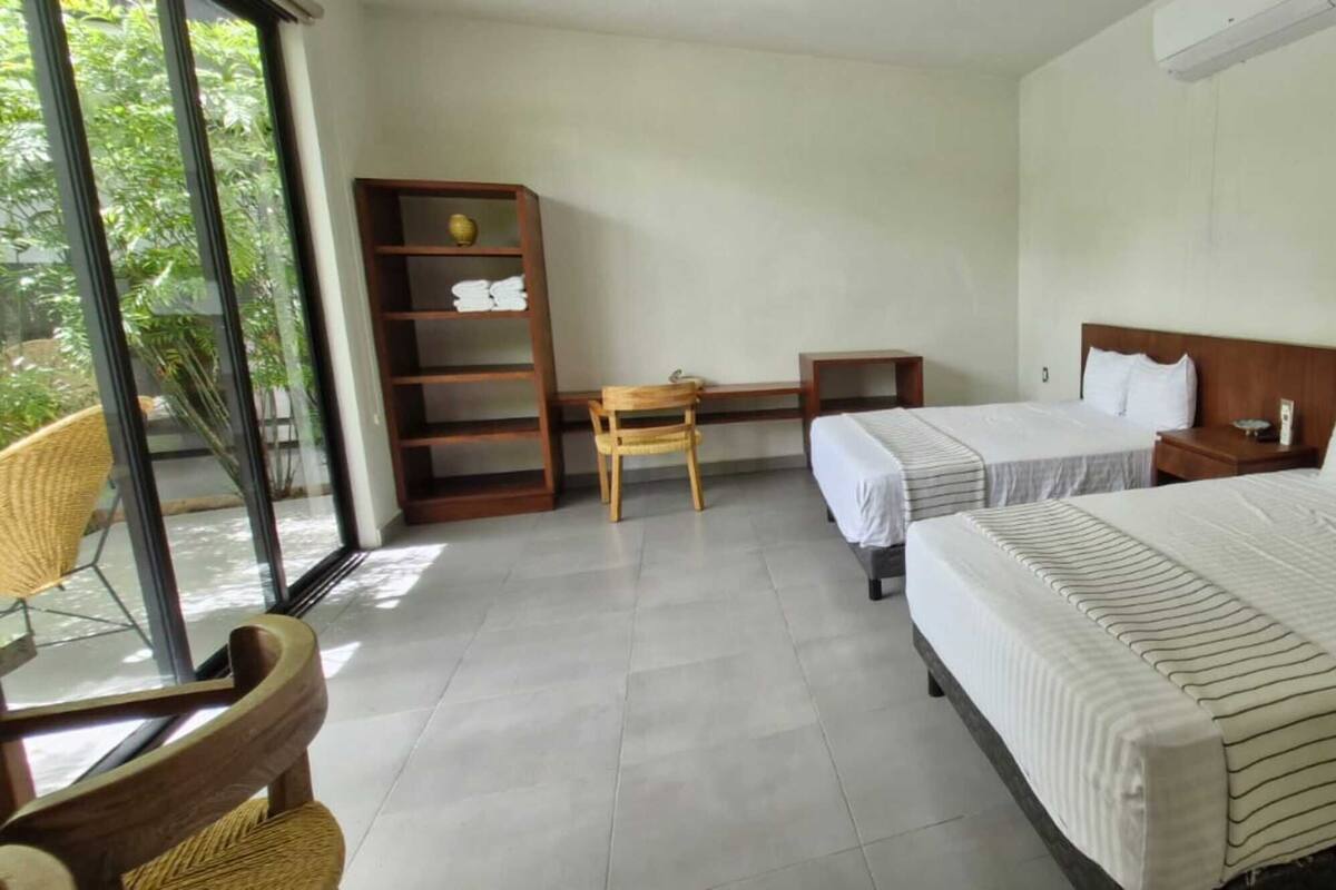 Habitación de hotel con dos camas, un escritorio de madera, una silla, una estantería y un ventanal grande.