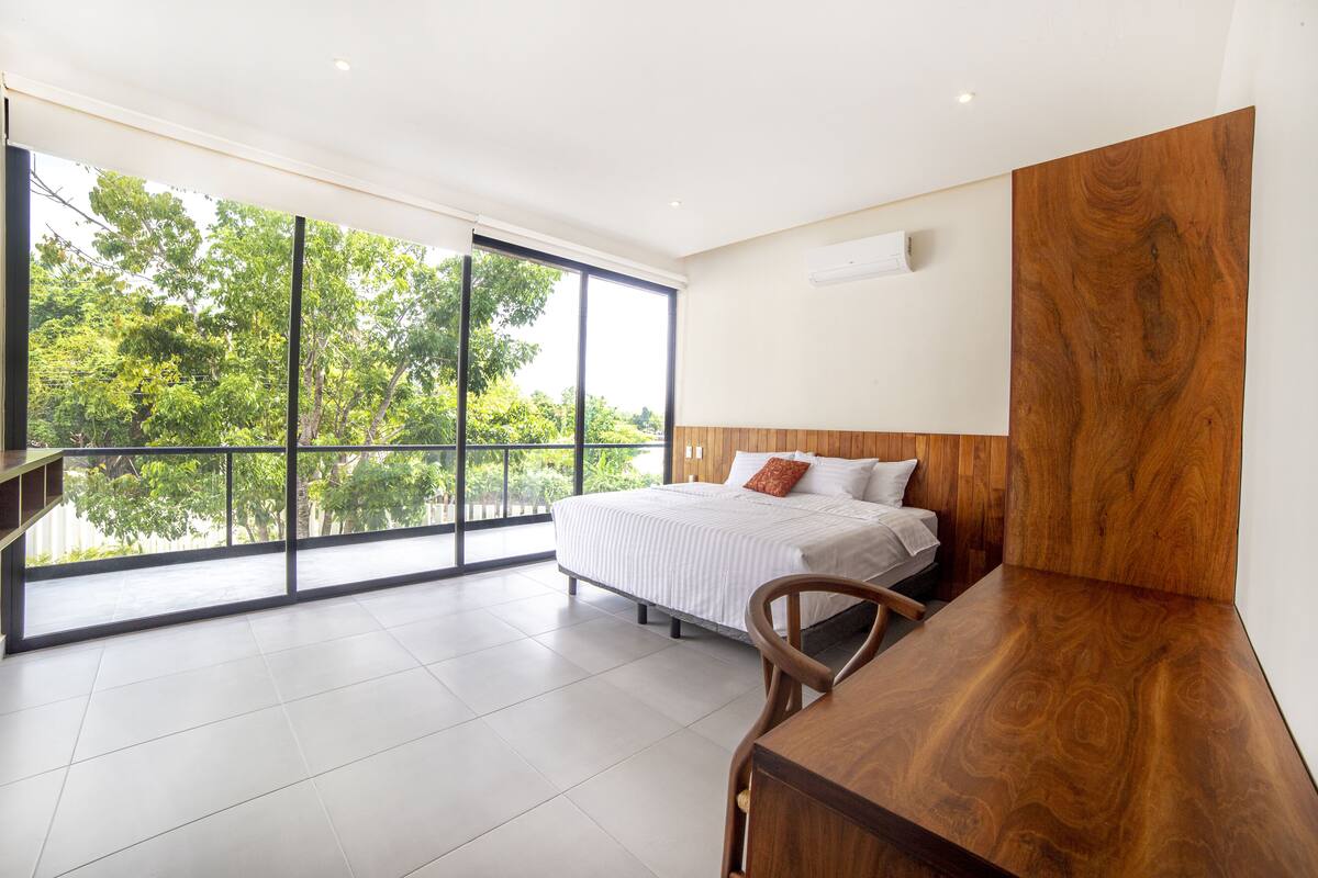 Un dormitorio moderno con un ventanal grande, una cama, un escritorio de madera y vistas a árboles.