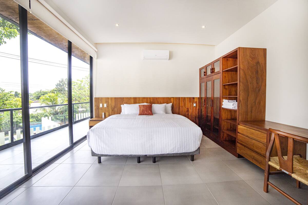Un dormitorio moderno con una cama grande, un armario de madera y vistas a la vegetación a través de amplias ventanas.