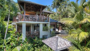 A two-story house with a balcony, surrounded by tropical vegetation.