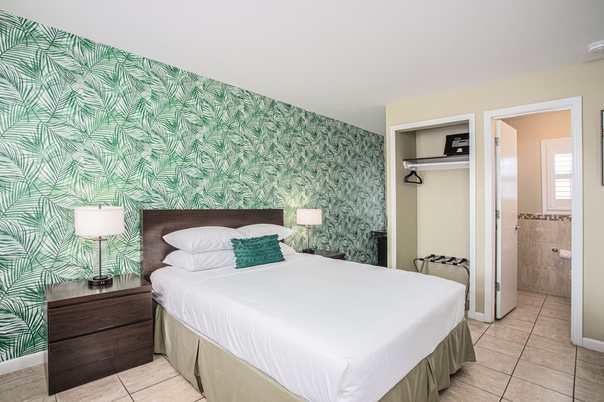 Un lit bien fait, avec du linge de lit blanc et un coussin vert en guise d’élément décoratif, une table de chevet avec une lampe, et un mur orné d’un motif végétal.