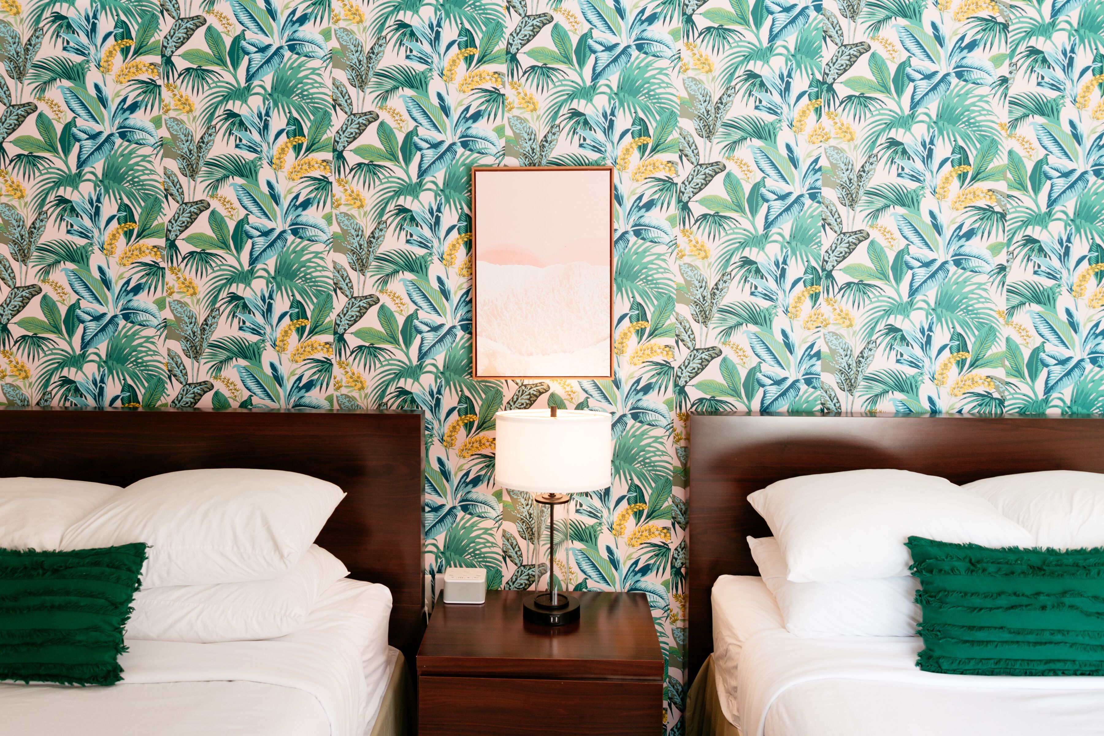 A hotel room with two beds, a nightstand with a lamp, and a tropical wallpaper.