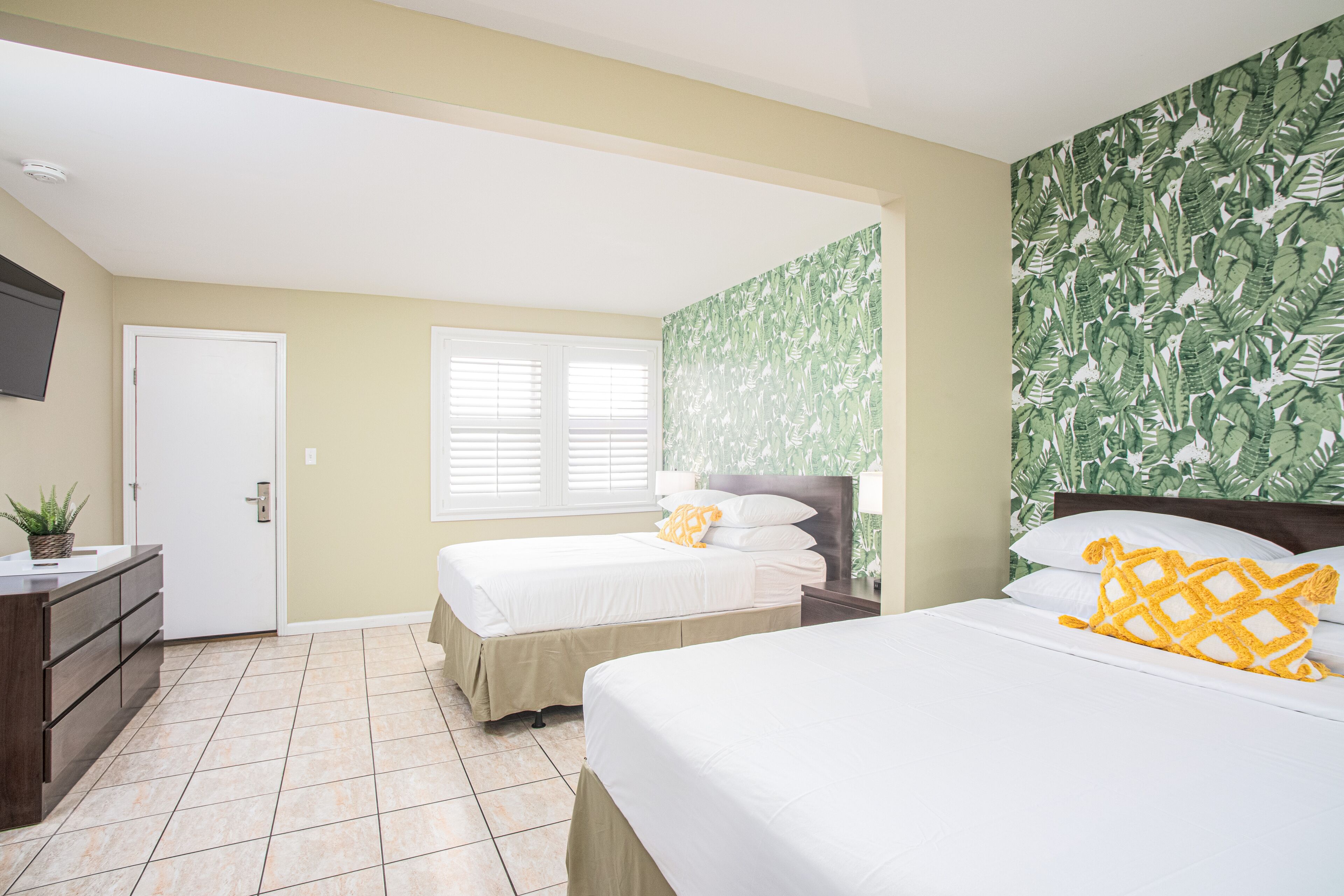 A hotel room with two beds, a small desk, a TV, and a window with blinds.