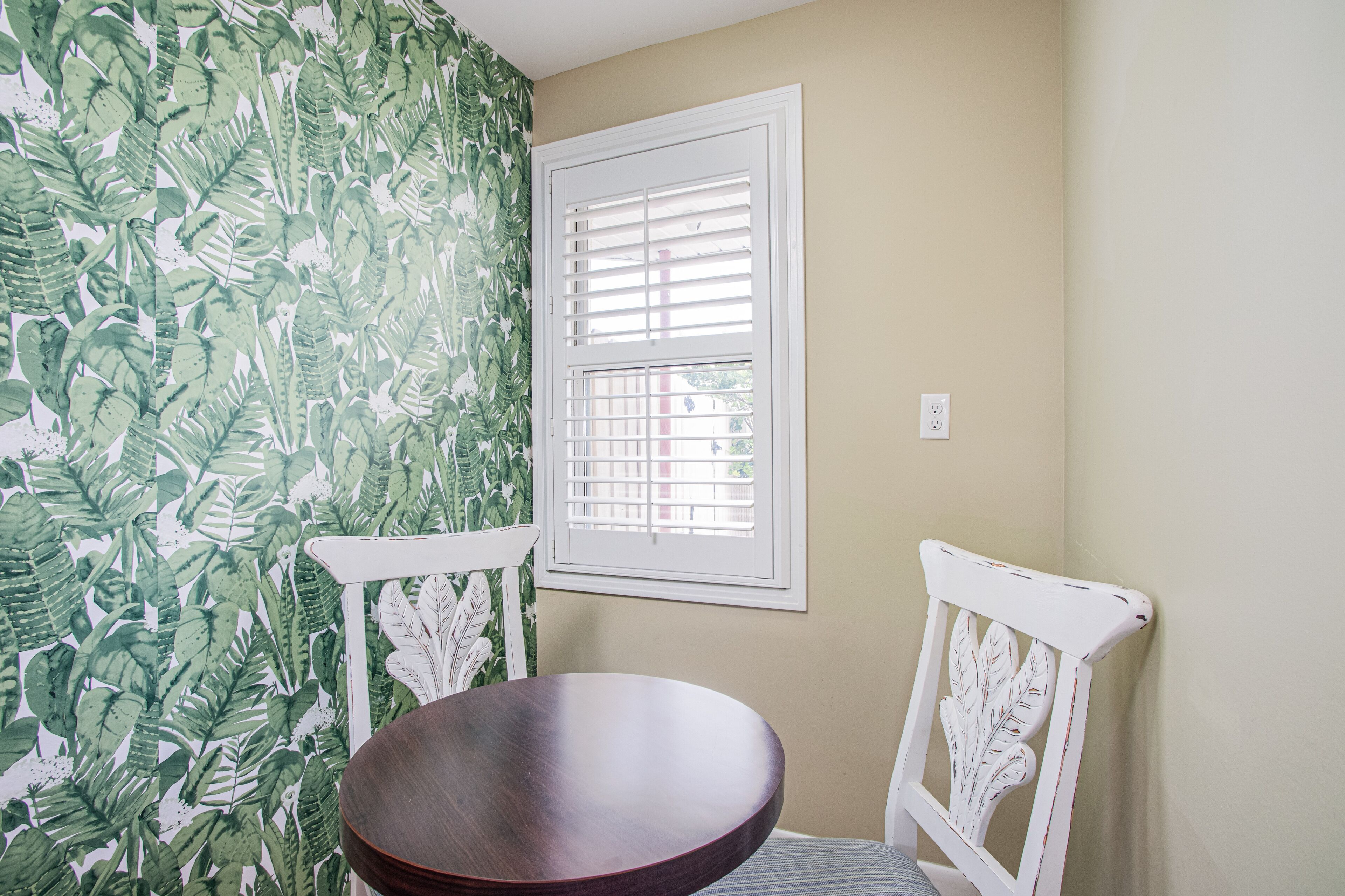 Una habitación con una mesa redonda de madera, dos sillas blancas, una ventana con contraventanas blancas y un papel tapiz con un estampado de hojas verdes.