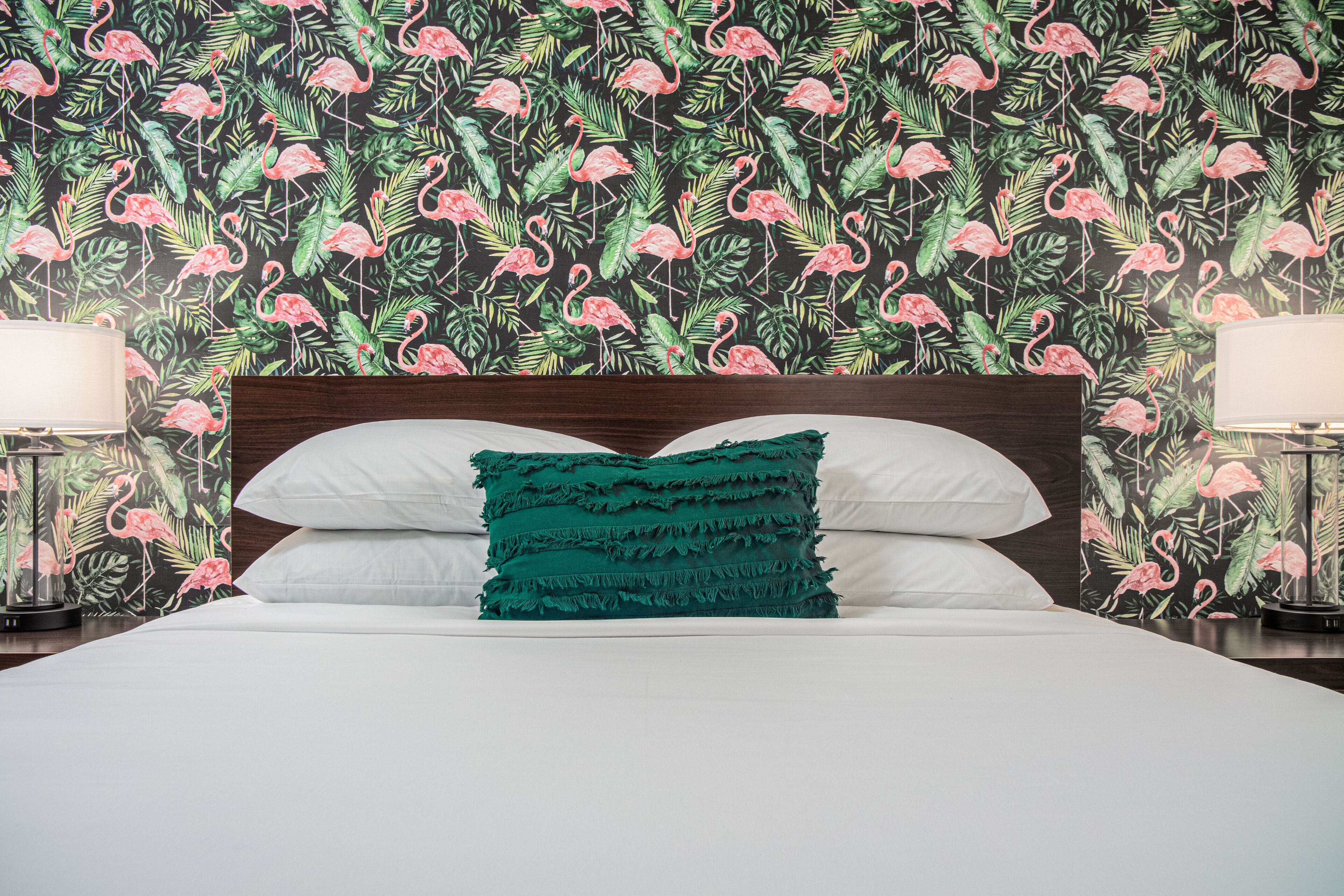 Una cama con sábanas blancas, un cojín de color aguamarina y un cabecero de madera. La habitación tiene un papel tapiz floral con flamencos rosados y hojas verdes.