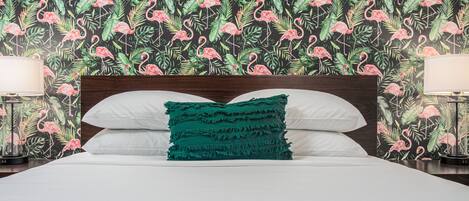 Una cama con sábanas blancas, un cojín de color aguamarina y un cabecero de madera. La habitación tiene un papel tapiz floral con flamencos rosados y hojas verdes.